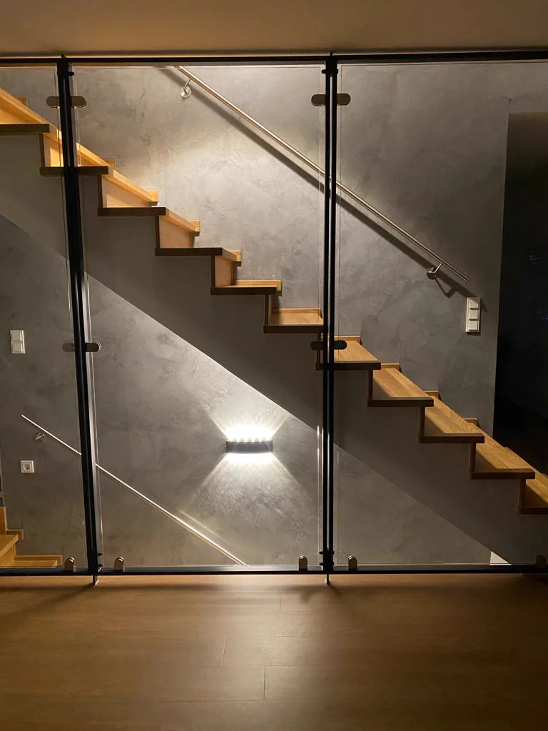Treppenaufgang mit Glasgeländer, Holzstufen und beleuchteter Wand in Spachteltechnik